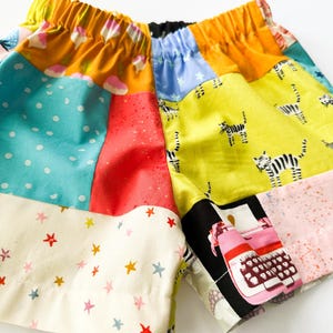May include: Colourful patchwork shorts with an elasticated waistband. The shorts feature various fabric patches with patterns like polka dots, stars, and a typewriter print. The colour scheme includes yellow, blue, red, and cream.