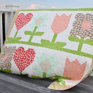 May include: A white quilt with a patchwork design featuring pink, red, and green flowers and hearts. The quilt is made with different patterned fabrics and is quilted with a simple stitch.