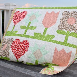 May include: A handmade quilt with a floral design. The quilt features large red hearts, pink tulips, and brown floral patterns on a white background. Green stems and a dotted green border complete the design. The quilt is draped over a wooden deck railing.