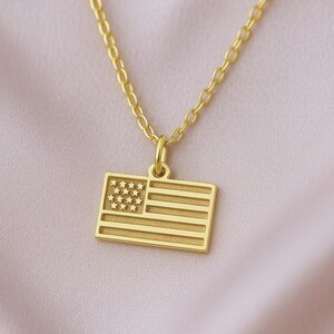 May include: A gold-toned necklace featuring a rectangular pendant shaped like the United States flag. The pendant displays a raised star pattern in the upper left corner and horizontal stripes across the rest of the surface, suspended from a delicate gold-toned chain.