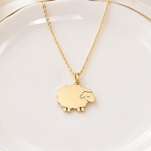 May include: A gold-coloured necklace featuring a sheep pendant. The pendant depicts a stylised sheep with a fluffy body and small legs. The chain is delicate and rests on a white plate.