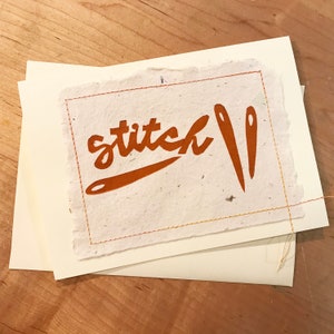 May include: A white card with a brown stitched border and the word "stitch" in brown lettering with two brown needles.