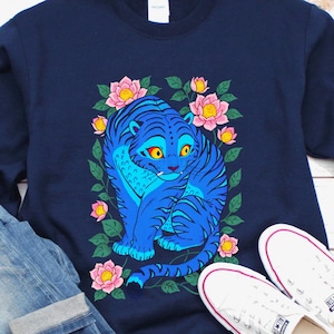 May include: Navy blue sweatshirt featuring a blue tiger design surrounded by pink flowers and green leaves. The tiger has yellow eyes and is set against a floral background. Light blue denim jeans and white sneakers complete the casual outfit.