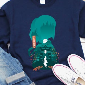 Op de afbeelding: Een marineblauw sweatshirt met een turquoise afbeelding van een silhouet en een Japanse landschap. Het sweatshirt wordt gecombineerd met een blauwe jeans en witte sneakers. Japanse tekst is zichtbaar.