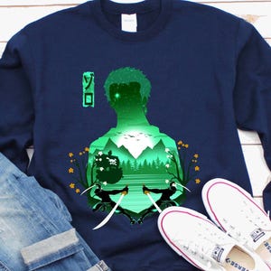 Może przedstawiać: Granatowa bluza z zielonym sylwetkowym wzorem osoby z mieczami, górami i japońskim tekstem. Obraz zawiera niebieskie dżinsy i białe płócienne buty.