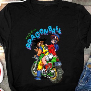 May include: A black t-shirt with a graphic of two anime characters riding a motorcycle. The characters are Goku and Bulma from the Dragon Ball series. The text "Dragon Ball" is written in a blue and white font above the characters. The text "ドラゴンボール" is written in Japanese below the characters.