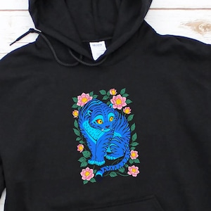 May include: Black hooded sweatshirt featuring a blue cat design with floral accents. The cat illustration is surrounded by pink and yellow flowers. The sweatshirt has a front pocket and a drawstring hood. The design is centered on the chest.