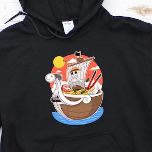 以下が含まれることがあります： ラーメンのボウルに入った漫画の海賊船のグラフィックがプリントされた黒いパーカー。船はドクロとクロスボーンの旗を持ち、ラーメンには箸とゆで卵が入っています。