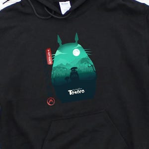 Op de afbeelding: Zwarte hoodie met een grafische print op de voorkant van Totoro in een boslandschap onder een volle maan. Het ontwerp gebruikt groen- en blauwtinten, met het woord "Totoro" eronder gedrukt.
