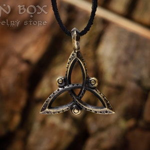 Könnte beinhalten: Eine goldfarbene Halskette mit einem keltischen Knoten-Anhänger und einer schwarzen Schnur. Der Anhänger ist ein stilisiertes dreifaches Knotendesign mit einer strukturierten Oberfläche.