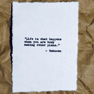 May include: A white rectangular paper with a quote printed in black ink: "Life is what happens when you are busy making other plans." The quote is attributed to "Unknown." The paper has a rough, textured edge and is set against a brown, crinkled background.