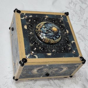 May include: A square wooden box featuring a dark blue celestial pattern with moons and stars. The lid displays a circular relief of a face profile with a sun and moon. The box is accented with gold-toned edges, small black beads, and clear rhinestones.