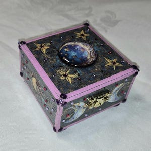 Puede incluir: Pequeña caja decorativa cuadrada con temática celestial. La caja presenta un fondo azul oscuro con estrellas doradas y un diseño de luna. Los bordes están pintados de rosa y adornados con gemas moradas. Un cierre dorado asegura la parte delantera.