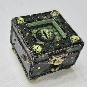 Puede incluir: Una caja de joyería cuadrada de color verde oscuro con tapa con bisagras y cierre de latón. La caja está decorada con múltiples adornos en forma de ojo verdes y amarillos y pequeñas gemas brillantes. La parte superior presenta una ventana transparente que muestra un gran diseño de ojo.
