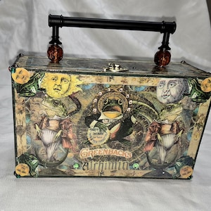 Op de afbeelding: Een decoratieve doos met een zwarte handgreep en decoratieve accenten. De doos heeft een collage-ontwerp met kikkers, zonnen, manen en de tekst "Greenbacks Alchimia". De doos heeft een vintage, grillige uitstraling.