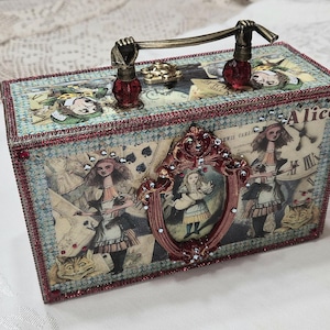 Op de afbeelding: Een rechthoekige decoratieve doos met Alice in Wonderland-afbeeldingen, waaronder speelkaartmotieven, een klok en de Cheshire Cat. De doos heeft een metalen handvat met rode kralen, een centraal rood ornament en een rode strass-afwerking.