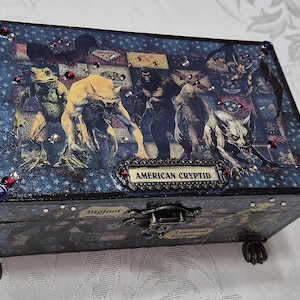 Puede incluir: Una caja rectangular de madera con un fondo azul oscuro con patrón de estrellas e ilustraciones de varios críptidos. Una placa central dice AMERICAN CRYPTID. La caja está decorada con pequeños diamantes de imitación rojos y transparentes y tiene patas en forma de garra.