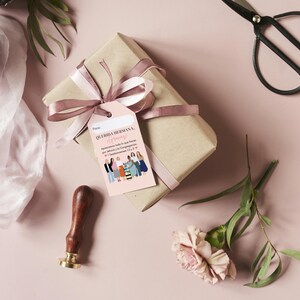 May include: A pink gift tag with a group of women illustrated on it. The tag reads "Querida Hermana, Happy Birthday!" and "Apreciamos todo lo que haces por nosotros. Te queremos mucho!"  The tag is tied to a brown gift box with a pink ribbon.