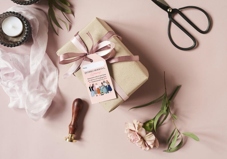 May include: A pink gift tag with a group of women illustrated on it. The tag reads "Querida Hermana, Happy Birthday!" and "Apreciamos todo lo que haces por nosotros. Te queremos mucho!"  The tag is tied to a brown gift box with a pink ribbon.