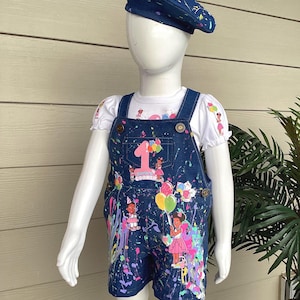 May include: A denim overall set for a child, featuring a beret and a short-sleeved shirt. The overalls are decorated with colorful paint splatters and a large number "1". The shirt has a matching design. The beret is navy blue with paint splatters.