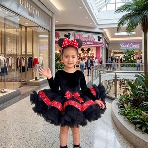 Può includere: Una bambina indossa un vestito di velluto nero con una gonna di tulle a strati, dettagli di nastro rosso e un grande fiocco. Indossa una fascia con orecchie di Minnie Mouse e scarpe Mary Jane rosse. L'outfit è un insieme giocoso a tema.