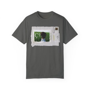 May include: A dark gray t-shirt with a vintage-style image of a young person looking out over a field of green grass. The image is framed with a white border and has a paper texture. The text "C+" is visible in the image.