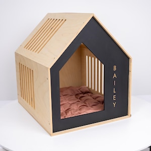 May include: A modern dog house in a house shape, constructed from light-colored wood with black trim. The front features a cutout with the name "BAILEY" in gold lettering. Inside is a pink quilted cushion. The roof and sides have ventilation slots.