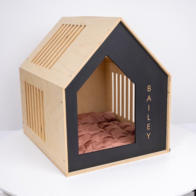May include: A modern dog house in a house shape, constructed from light-colored wood with black trim. The front features a cutout with the name "BAILEY" in gold lettering. Inside is a pink quilted cushion. The roof and sides have ventilation slots.