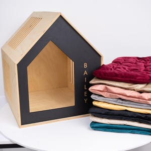 May include: A wooden dog house with a black trim and the name "BAILEY" in gold lettering. A stack of colorful pet bed cushions in various colors, including burgundy, tan, pink, and teal, sits next to the house.