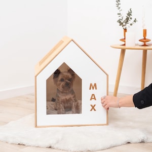 May include: A modern dog house in a house shape with a white front and natural wood trim. The name "MAX" is displayed on the side. A small brown dog sits inside the house on a soft bed.