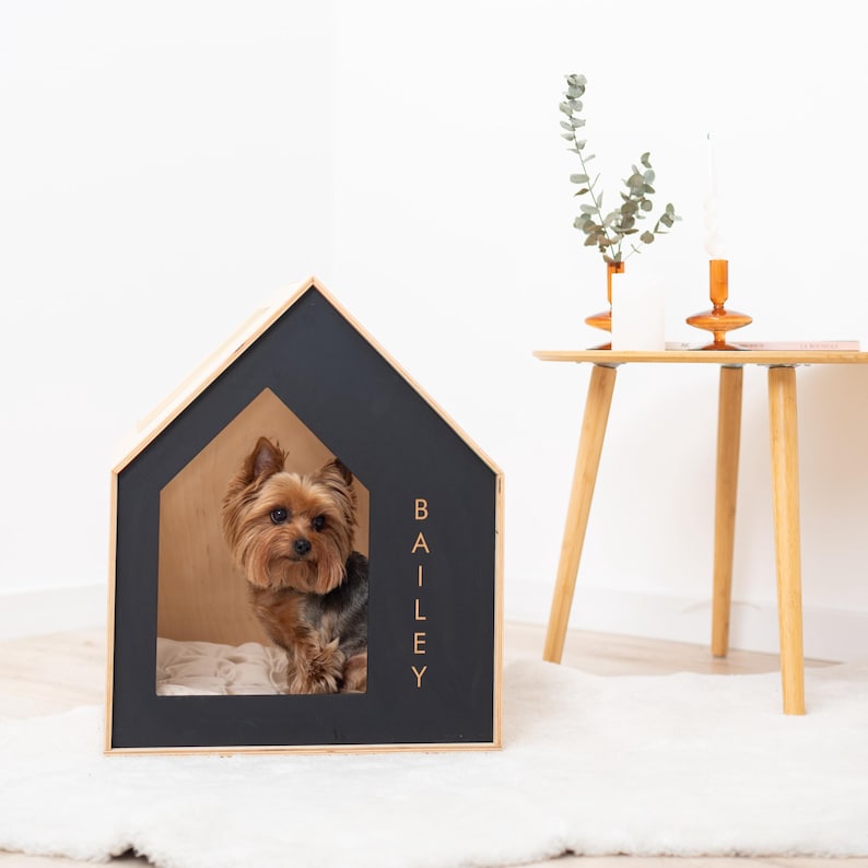 May include: A wooden dog house with a black exterior and natural wood trim. The house has a house-shaped design with the name "BAILEY" printed vertically on the side. A small dog is inside the house, sitting on a white cushion.