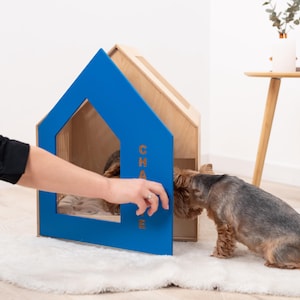 May include: A wooden dog house with a blue frame and the name "CHASE" in orange letters. The dog house has a house-like shape. A small dog is sniffing the entrance of the dog house.
