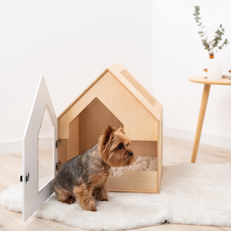 May include: A small dog sits near a wooden dog house with a white door. The house is light brown with a house-shaped design and a window. The door is open, revealing the interior. The dog is brown and gray.