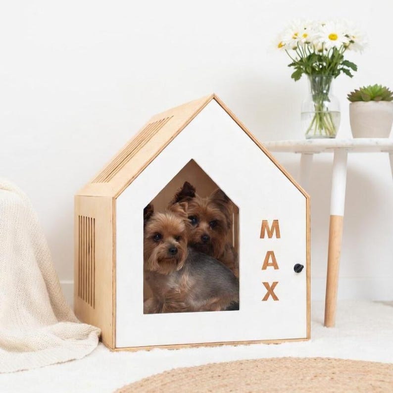 May include: A wooden dog house with a white front and a natural wood frame. Two small dogs are inside the house. The name "MAX" is printed on the side. A vase of white flowers and a small succulent plant are in the background.