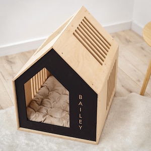 May include: A wooden dog house with a house-shaped design. The frame is light-colored wood with black accents around the entrance. The name "BAILEY" is printed vertically on the side. A beige cushion is inside. The roof has ventilation slats.
