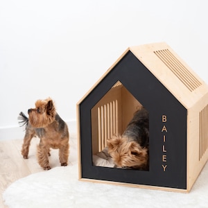 May include: A wooden dog house with a black trim and the name "BAILEY" in gold lettering. A small dog is inside the house, and another dog stands nearby. The house has a modern design with a slatted roof and side panels.