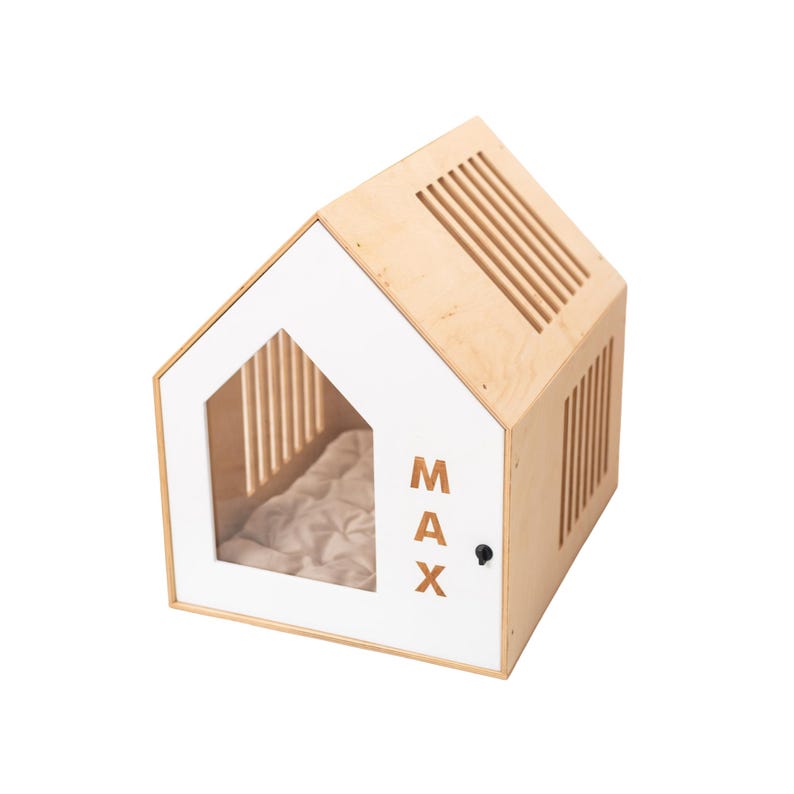 May include: A wooden dog house with a white front and a slanted roof. The front features a window and the name "MAX" in brown letters. The house has ventilation slats and a small black door handle. Inside is a beige cushion.