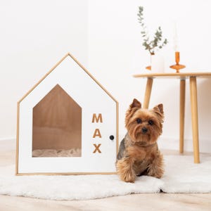 May include: A white dog house with a wooden frame and a house-shaped opening. The name "MAX" is printed on the side. A small dog with brown and tan fur sits in front of the dog house on a white rug.