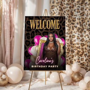 May include: A welcome sign on a gold easel with the text "WELCOME" in gold glitter. The sign features a woman in a leopard print dress, surrounded by pink and white balloons. The text "Carolina's BIRTHDAY PARTY" is also displayed.