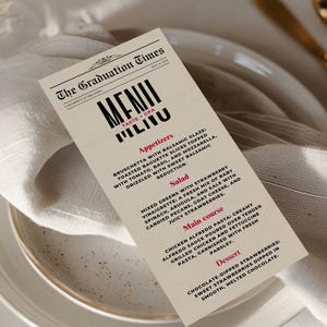 May include: A cream-coloured paper menu titled The Graduation Times, featuring black and red text listing appetizers, salad, main course, and dessert options. The menu is placed on a white plate with a folded white napkin and gold-toned cutlery.