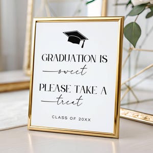 May include: A framed graduation sign with a gold border. The sign reads "Graduation is sweet, Please take a treat" with a graduation cap graphic. The text "Class of 20XX" is at the bottom. The sign is on a white surface.
