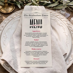Op de afbeelding: Een rechthoekig papieren menu met de titel The Graduation Times met zwarte en roze tekst met een lijst van voorgerechten, salades, hoofdgerechten en desserts. Het menu ligt in het midden op een wit linnen servet op een wit keramieken bord.