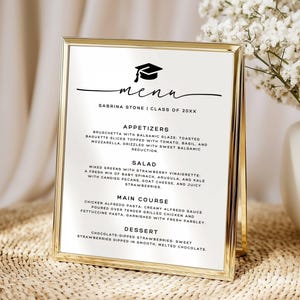May include: A gold-framed vertical menu sign featuring a black graduation cap icon and the text "MENU" followed by "SABRINA STONE | CLASS OF 20XX". The menu lists appetizers, salad, main course, and dessert options in black sans-serif font.