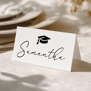 May include: A white place card with the name "Samantha" in elegant black script, topped with a black graduation cap icon. The card is folded and set on a textured, off-white surface, suggesting a graduation or celebratory event.
