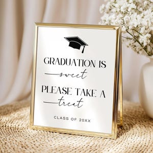 Könnte beinhalten: Ein goldgerahmtes Schild mit dem Text "GRADUATION IS sweet PLEASE TAKE A treat CLASS OF 20XX" und einer Abschlusskappen-Ikone. Das Schild steht auf einer gewebten Oberfläche, mit weißen Blumen in einer Vase im Hintergrund.