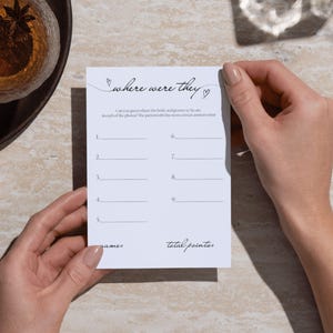 May include: A white card with the text "where were they" in cursive script, held by two hands. The card has lines for answers and the words "games" and "total points". A pen and a drink are in the background.