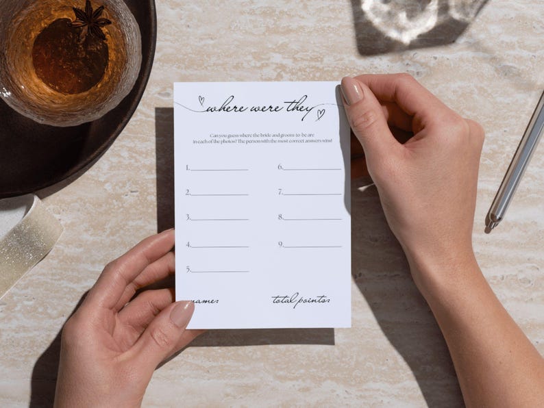 May include: A white card with the text "where were they" in cursive script, held by two hands. The card has lines for answers and the words "games" and "total points". A pen and a drink are in the background.