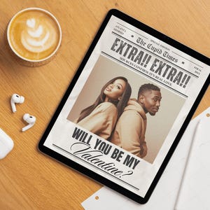 May include: A digital tablet displaying a newspaper-style graphic with the headline "EXTRA!! EXTRA!!" and the text "WILL YOU BE MY Valentine?" A latte and wireless earbuds are on a wooden surface.