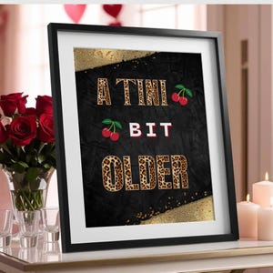May include: A black and gold framed print with the text "A TINI BIT OLDER" in leopard print font with red cherries on either side of the text.