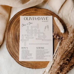 K&ouml;nnte beinhalten: Ein Hochzeits-Kreuzwortr&auml;tsel mit den Namen "OLIVIA + DAVE" und dem Datum "05.09.202X" oben. Das R&auml;tsel enth&auml;lt Hinweise auf Braut und Br&auml;utigam, mit Champagnergl&auml;sern illustriert. Das R&auml;tsel befindet sich auf hellem Papier.